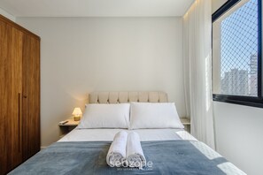 Una cama bien hecha con sábanas blancas y toallas enrolladas, un armario de madera, una mesita de noche con una lámpara, y una ventana con vistas a edificios.