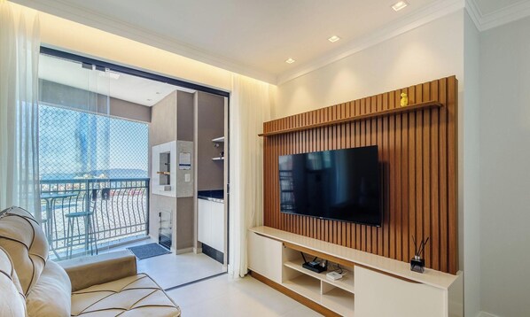 Una sala de estar moderna con un televisor de pantalla plana grande montado en una pared de paneles de madera, un sofá y una puerta de cristal que da a un balcón con mobiliario exterior.