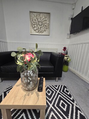 A living room with a black leather sofa, a wooden coffee table with a remote control, a patterned rug, and a wall-mounted TV. - Private & Charming Annex. Wi-Fi, Free parking, 15 min to Swindon Designer shops. (Highworth)