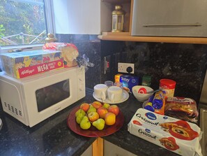 A kitchen counter with a microwave, a plate of apples, a bowl of bread, and various packaged food items. - Private & Charming Annex. Wi-Fi, Free parking, 15 min to Swindon Designer shops. (Highworth)