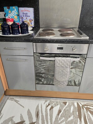 Eine Küche mit Herd, Backofen und einer Arbeitsfläche, auf der Gläser mit den Aufschriften 'TEE', 'KAFFEE' und 'ZUCKER' stehen.