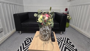 A living room with a black leather sofa, a wooden coffee table with a vase of flowers, a remote control, and a wall-mounted TV. - Private & Charming Annex. Wi-Fi, Free parking, 15 min to Swindon Designer shops. (Highworth)