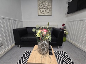 A living room with a black leather sofa, a wooden coffee table with a vase of flowers, a remote control, and a wall-mounted TV. - Private & Charming Annex. Wi-Fi, Free parking, 15 min to Swindon Designer shops. (Highworth)