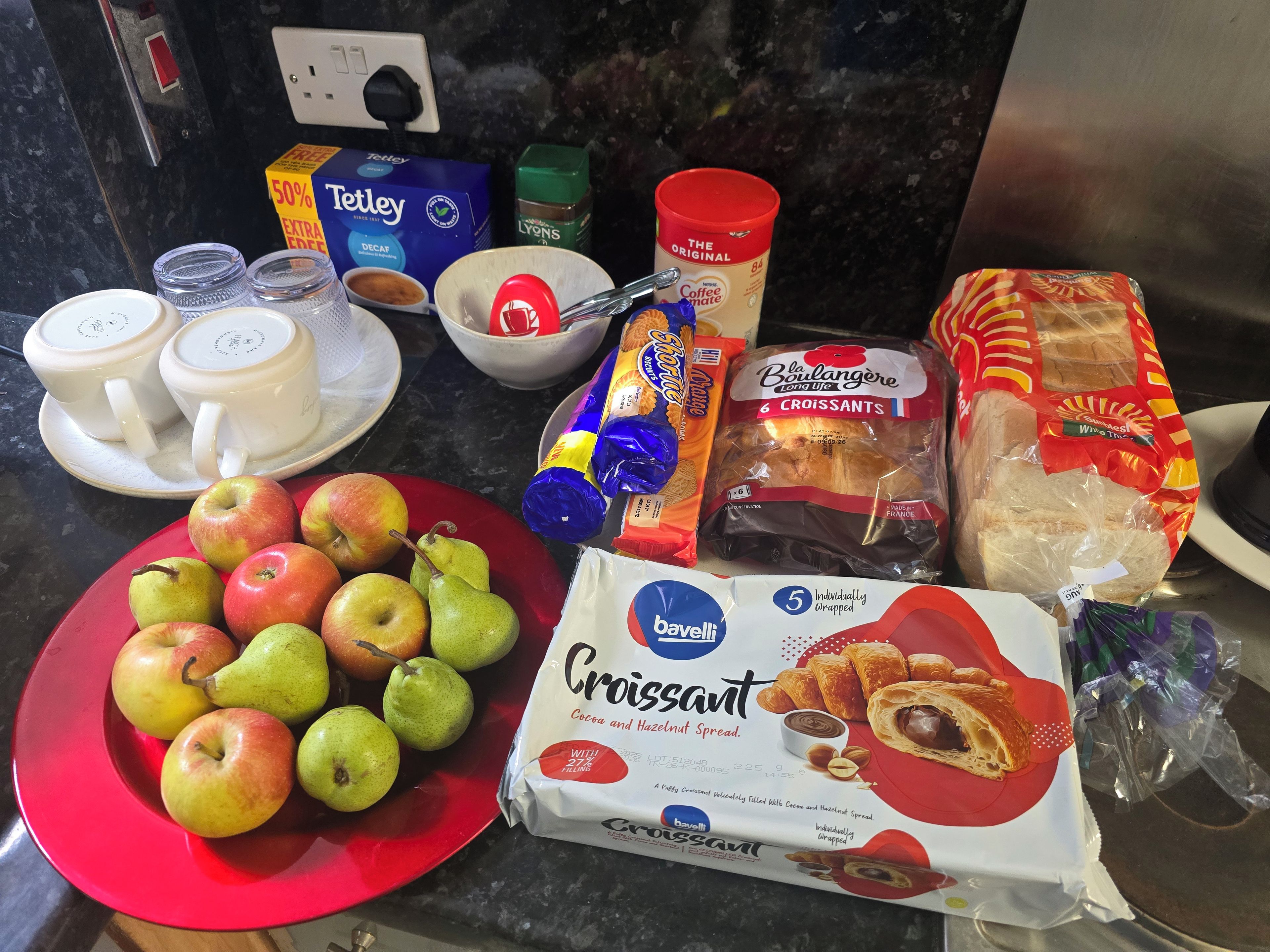 A kitchen counter with various food items including apples, pears, bread, and a package of croissants.