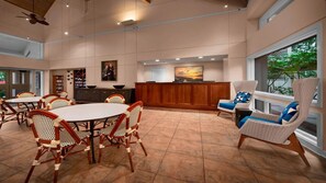Un espacio interior amplio con una mesa de recepción, una zona de comedor con mesas redondas y sillas de mimbre, y un gran ventanal con vistas a zonas verdes.