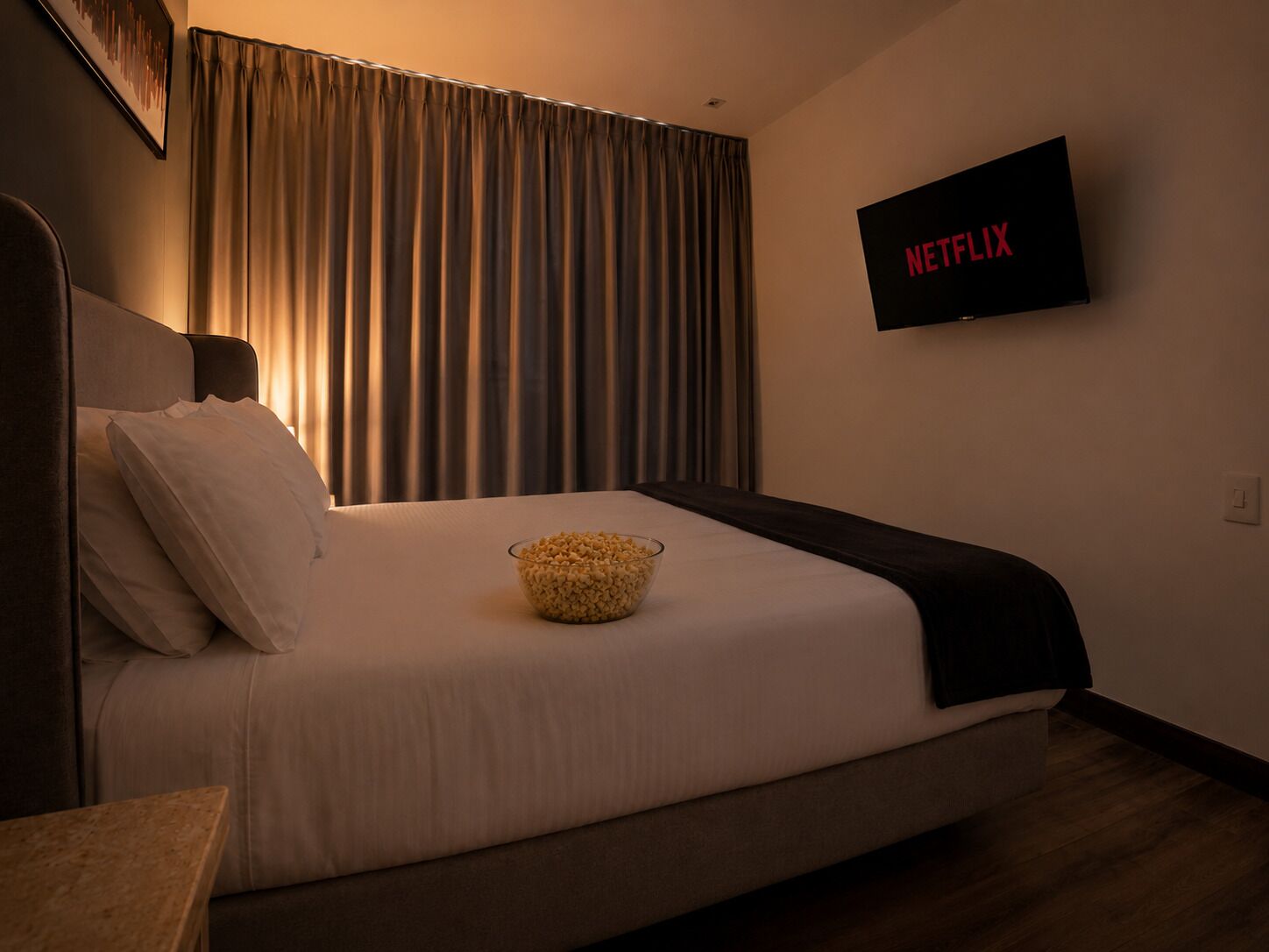 A hotel room with a bed, a bowl of popcorn, a TV displaying 'NETFLIX', and a curtain.
