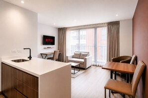 Habitación de hotel moderna con kitchenette, televisor de pantalla plana, sofá, mesa de comedor y un amplio ventanal con cortinas.