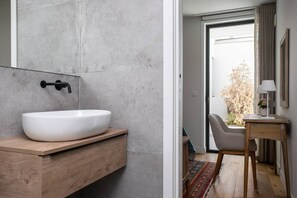 Ein modernes Badezimmer mit einem großen weißen Waschbecken, einem Holzkosmetikschrank und Blick ins Freie durch eine Glastür.
