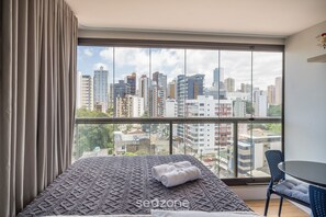 Ein Hotelzimmer mit großem Fenster und Blick auf die Stadt, einem Bett mit gemustertem Überwurf sowie einem kleinen Tisch mit Stuhl.