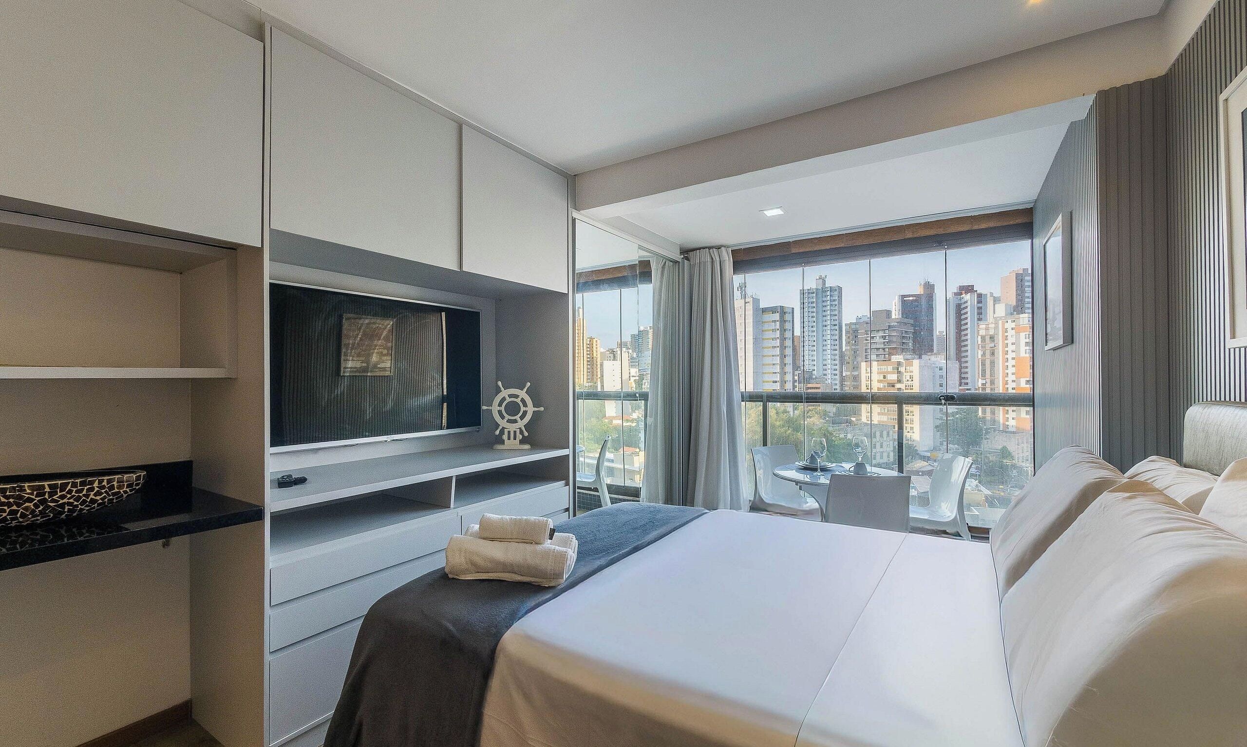 Een hotelkamer met een bed, een televisie, uitzicht op de stad en een raam met gordijnen.