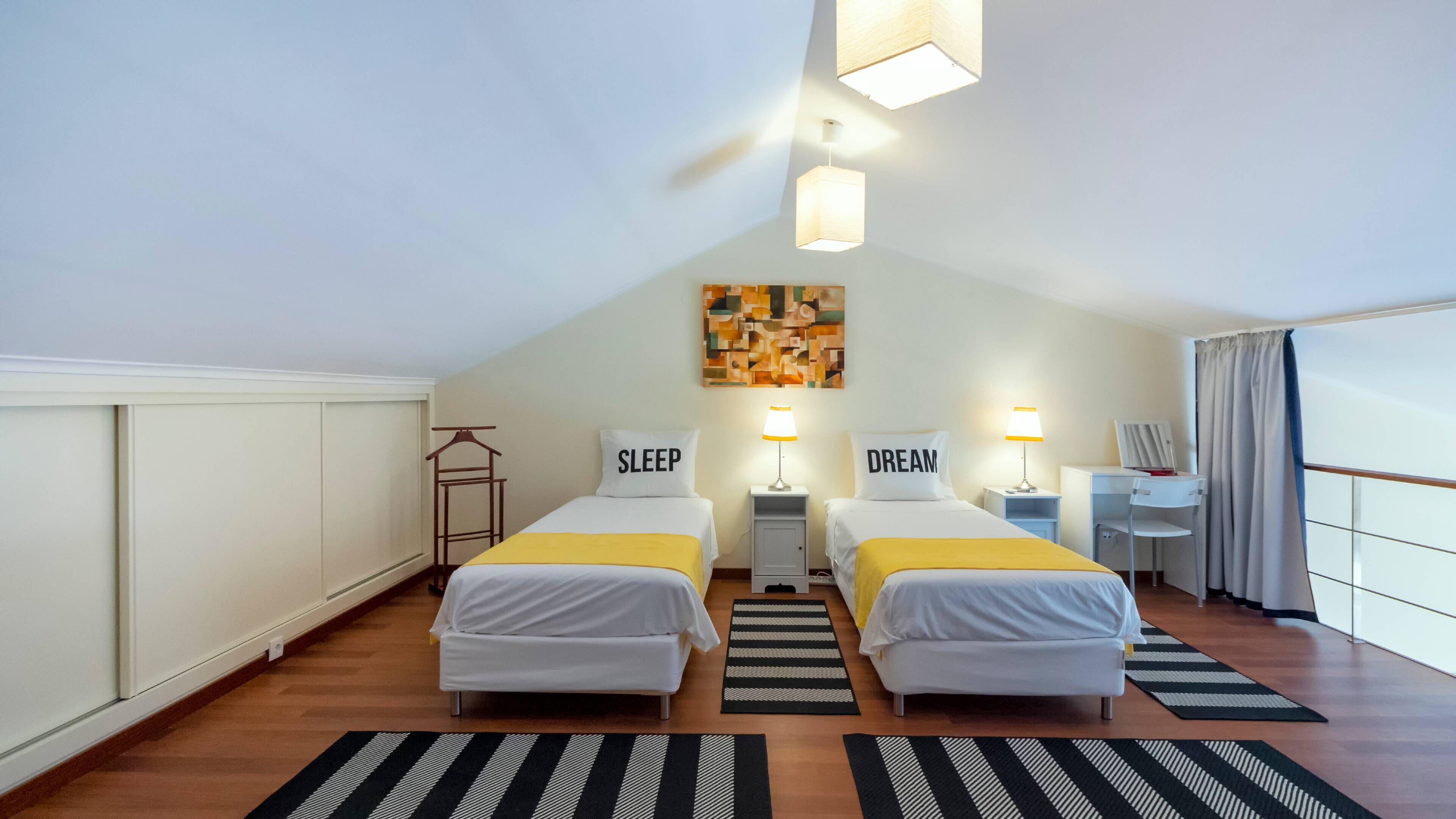 A hotel room with two beds, a desk, a chair, a hanging light fixture, and a painting on the wall.