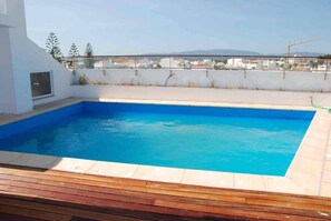 A rectangular swimming pool with a wooden deck in the foreground, surrounded by a white wall, with a view of buildings and mountains in the background. - The Sea Terrace - Rooftop Beach Duplex - Centre (Portimão)
