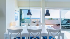 Un espace repas comprenant une table et des chaises, de grandes fenêtres offrant une vue sur la mer et des luminaires suspendus.