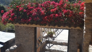 A stone structure with a wooden roof, a wheel, and a garden with red flowers.