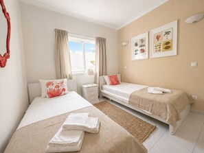 Une chambre d’hôtel avec deux lits, une fenêtre avec des rideaux, un mur orné d’illustrations botaniques encadrées et une table de chevet avec une lampe.