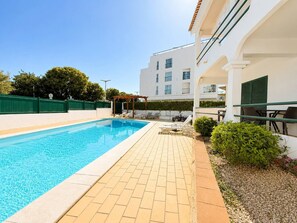 Une piscine avec une terrasse carrelée, des sièges extérieurs et un bâtiment doté de fenêtres, avec une clôture verte en arrière-plan.