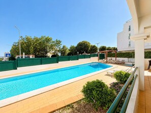 A rectangular swimming pool with a tiled deck, surrounded by a green fence, and a covered seating area with lounge chairs. - The Sea Breeze - Bright Beach Apartment - Centre (Portimão)
