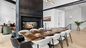 A modern dining area with a long wooden table set for six, surrounded by white upholstered chairs. A fireplace is visible in the background, and the room has a high ceiling with exposed beams. - EagleVail Designer Retreat | Sleeps 16 (Vail)