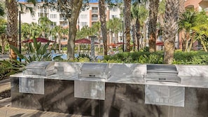 Un espace barbecue extérieur doté de plusieurs grils, entouré de palmiers et avec une piscine en arrière-plan.