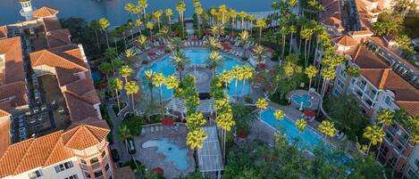 An aerial view of a resort with multiple swimming pools surrounded by palm trees and a waterfront.