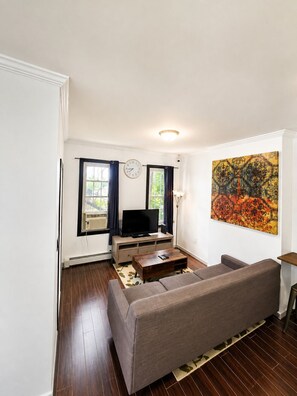 Ein Wohnzimmer mit einer Couch, einem Fernseher auf einem Schrank, einem Bild an der Wand und einem Fenster mit Vorhängen.