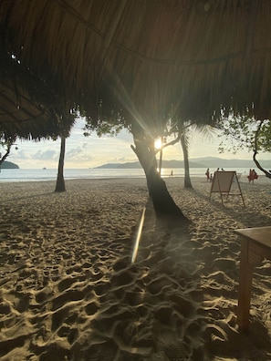 A beach scene with a thatched roof structure, a sandy beach, and a calm sea. - Villa Violeta | Ocean Views Above Playa La Ropa (Zihuatanejo)