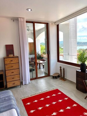 A modern bedroom with a sliding glass door leading to a balcony, a wooden dresser, a red patterned rug, and a view of the outdoors. - Villa Violeta | Ocean Views Above Playa La Ropa (Zihuatanejo)