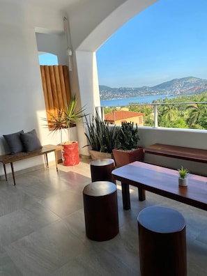 A modern balcony with a wooden bench, potted plants, and a view of a coastal landscape. - Villa Violeta | Ocean Views Above Playa La Ropa (Zihuatanejo)