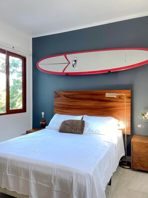 Een slaapkamer met een bed, een nachtkastje, een surfboard decoratie aan de muur en een raam met uitzicht op bomen.