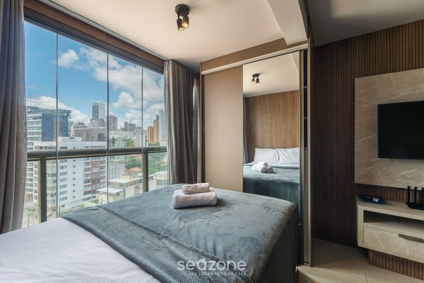 Hotelkamer met groot raam en uitzicht op de stad, een bed met grijze plaid, een flatscreen-televisie aan de muur en een houten paneelwand.