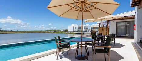 A poolside area with a table and chairs, a large umbrella, and a view of a body of water and buildings in the background.