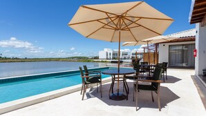 A poolside area with a table and chairs, a large umbrella, and a view of a body of water and buildings in the background. - House by Lagoon, Near Beach Cdl0007 (Maraú)