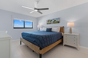 A bedroom with a bed, a nightstand, a lamp, a ceiling fan, a window, and a painting on the wall. - Sunset Condo #502 (Fort Myers Beach)