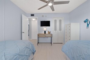 Een slaapkamer met twee bedden, een plafondventilator, een televisie, een bureau met lamp en een kledingkast.