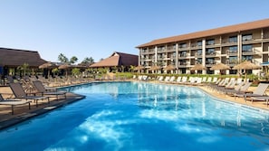 Ein Poolbereich mit Liegestühlen und Sonnenschirmen, ein großes Gebäude mit mehreren Balkonen und einem strahlend blauen Himmel.