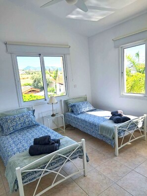 A room with two beds, a ceiling fan, and large windows with a view of greenery and buildings. - Villa Ella - peaceful detached villa close to beach and mountains - Sleeps 6 (Argaka)