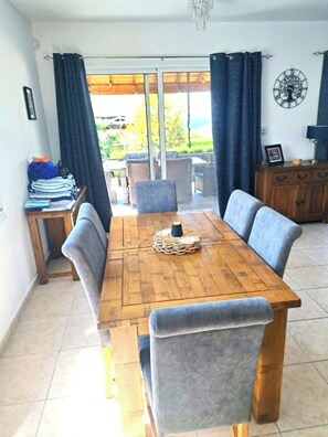A dining area with a wooden table and cushioned chairs, a small side table with a lamp, and a view through a glass door to the outside. - Villa Ella - peaceful detached villa close to beach and mountains - Sleeps 6 (Argaka)