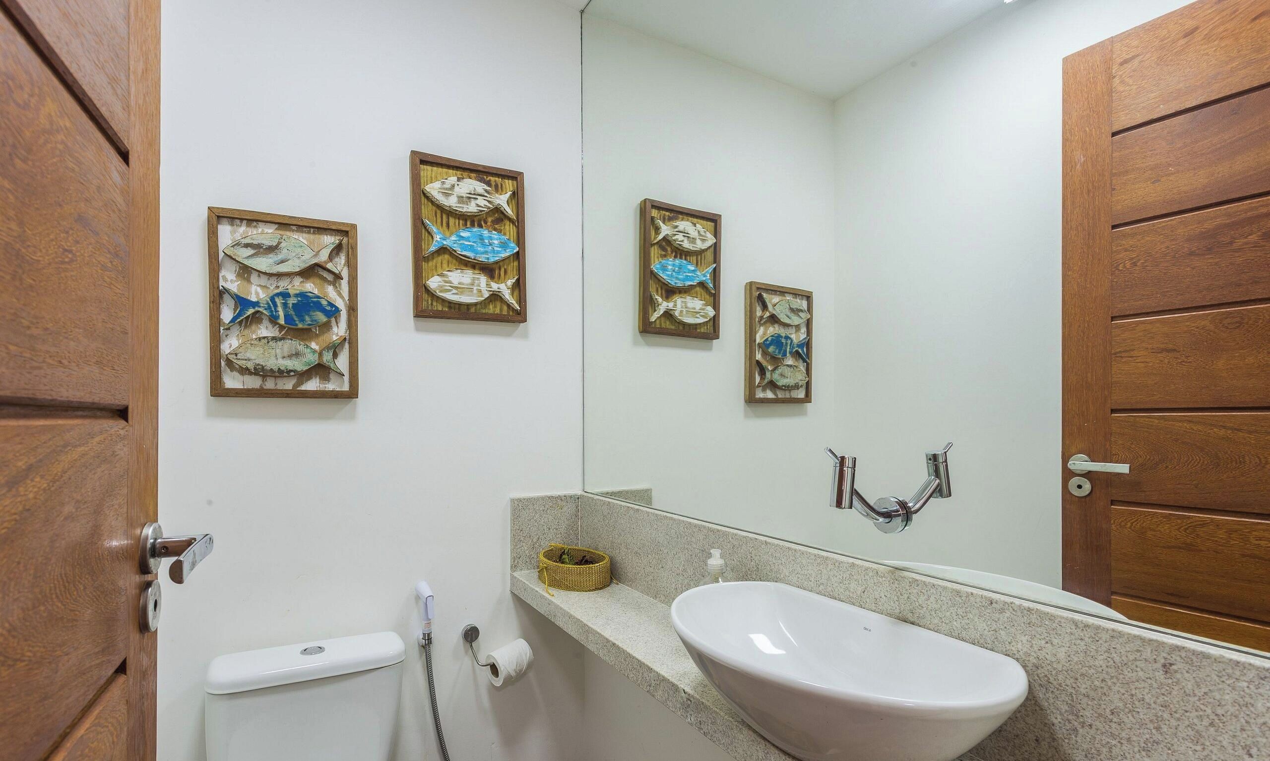 Une salle de bain avec une porte en bois, un lavabo blanc, un miroir et trois œuvres d’art mural représentant des poissons, encadrées.