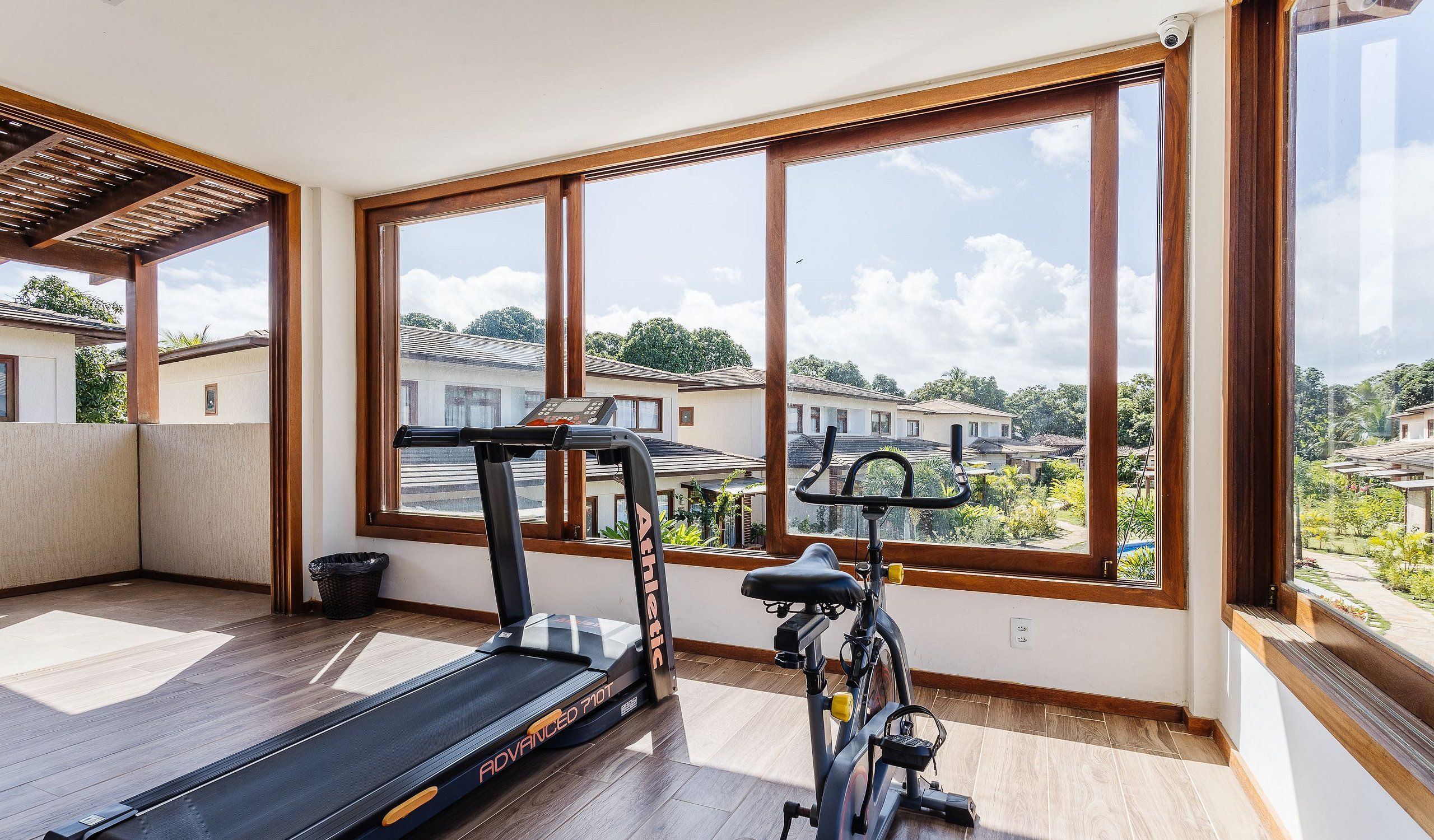Varanda espaçosa com esteira e bicicleta ergométrica, grandes janelas com vista para uma área residencial.
