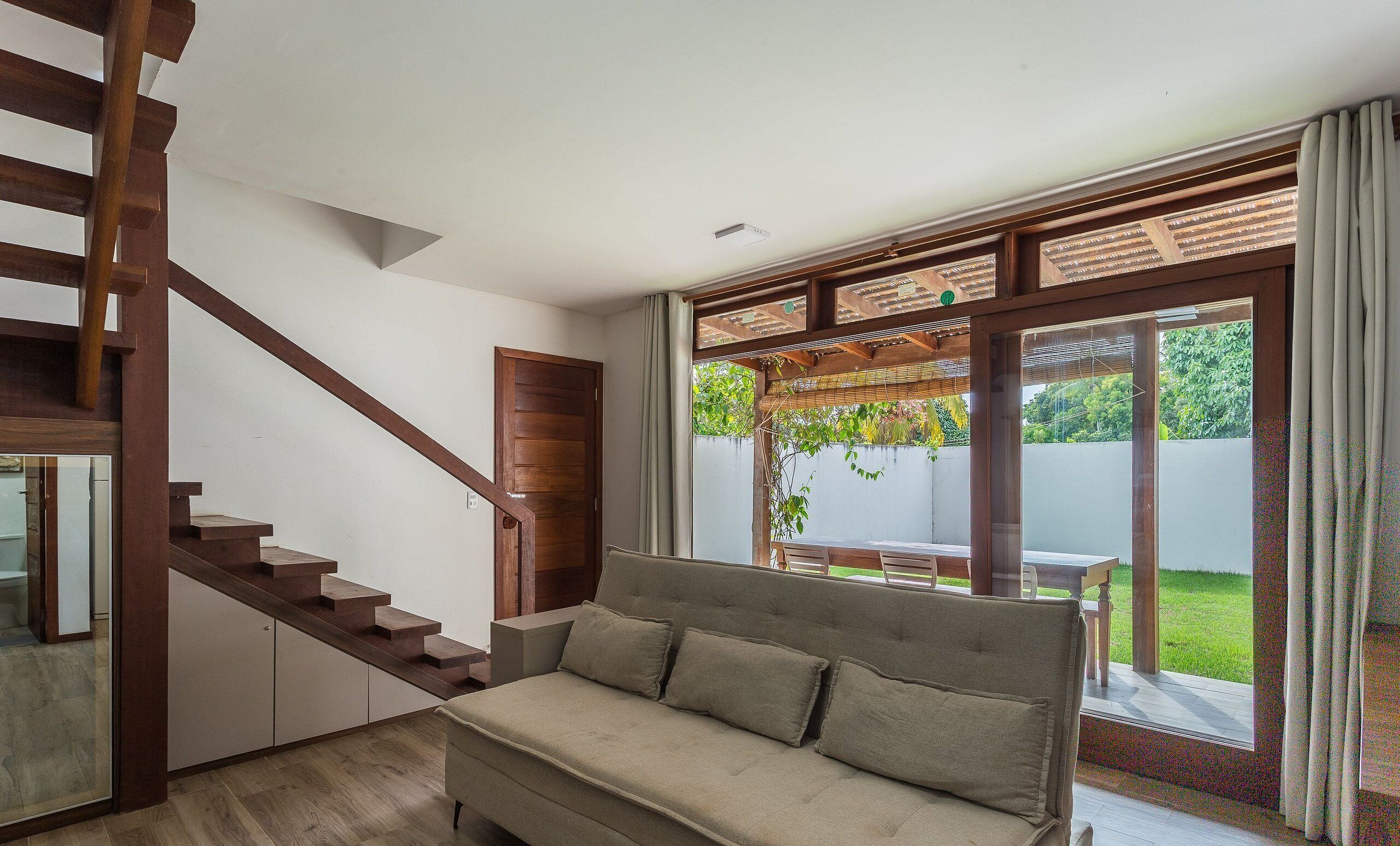 A modern living room with a wooden staircase, a sofa, and a large window with a view of greenery.