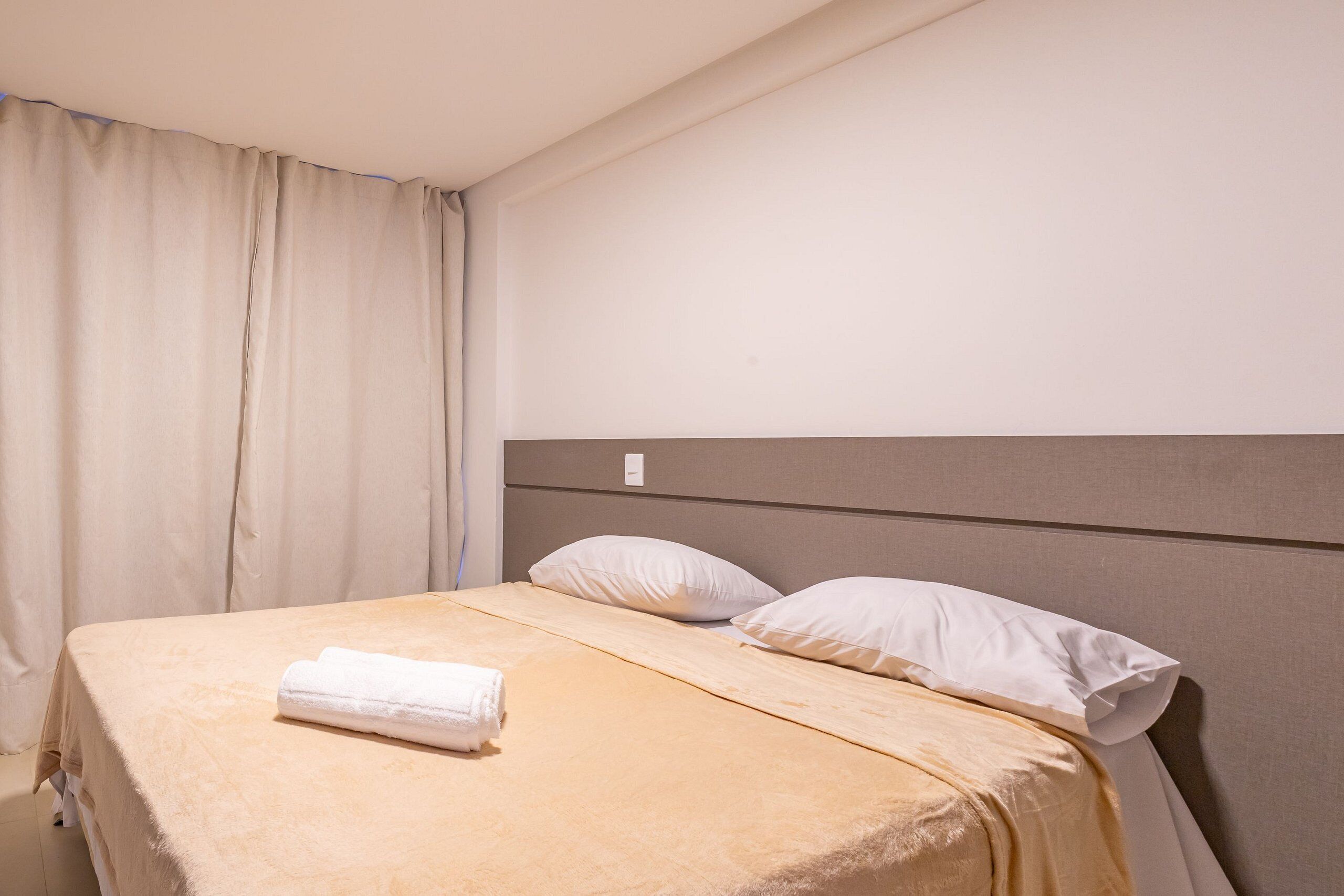A neatly made bed with white linens and a beige blanket, a headboard, and a window with curtains.