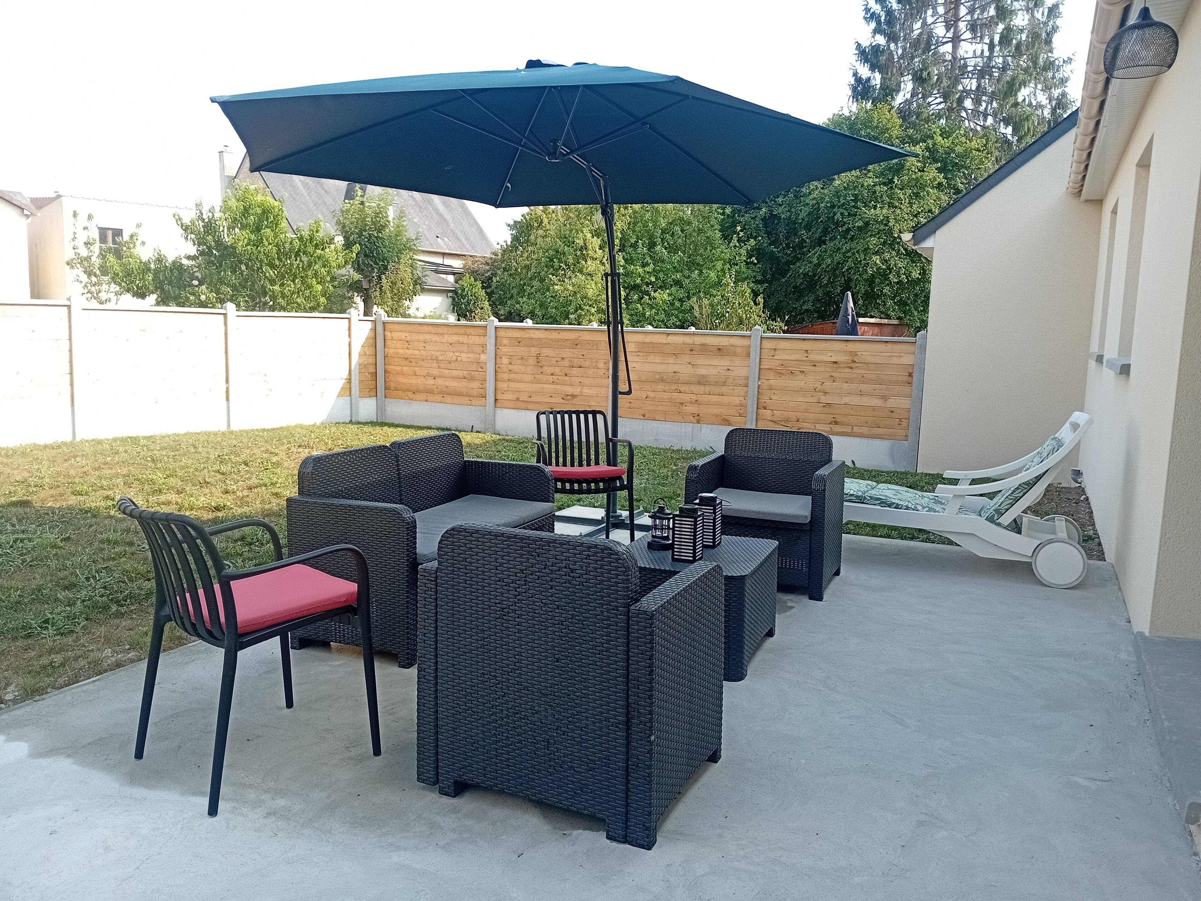 Une terrasse extérieure avec des meubles en osier, un parasol bleu et une chaise longue blanche.