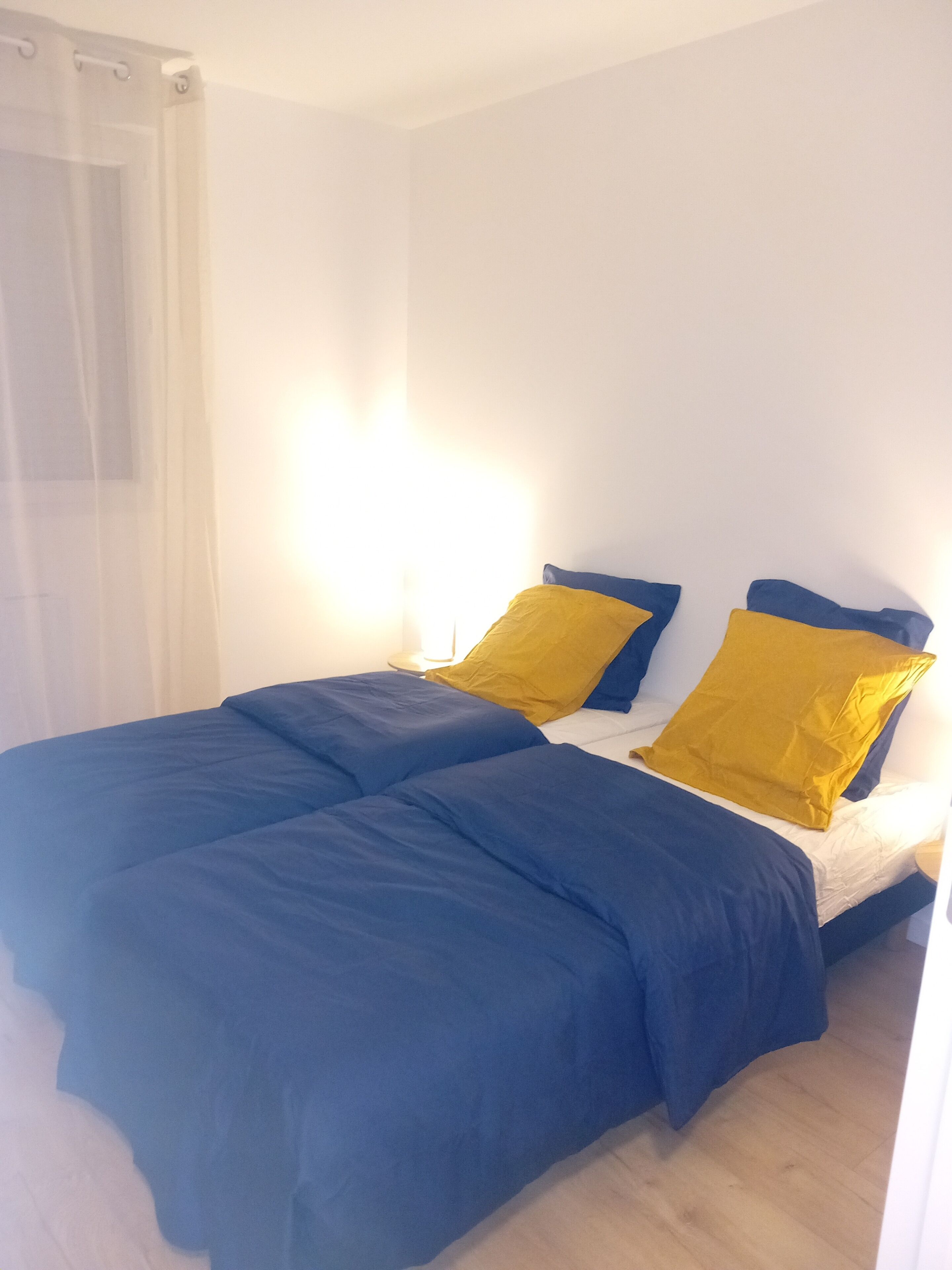 Un letto con biancheria azzurra e due cuscini gialli, una tenda bianca e una fonte di luce a parete.