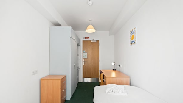 A hotel room with a bed, a desk, a wardrobe, a door, and a wall-mounted light fixture.