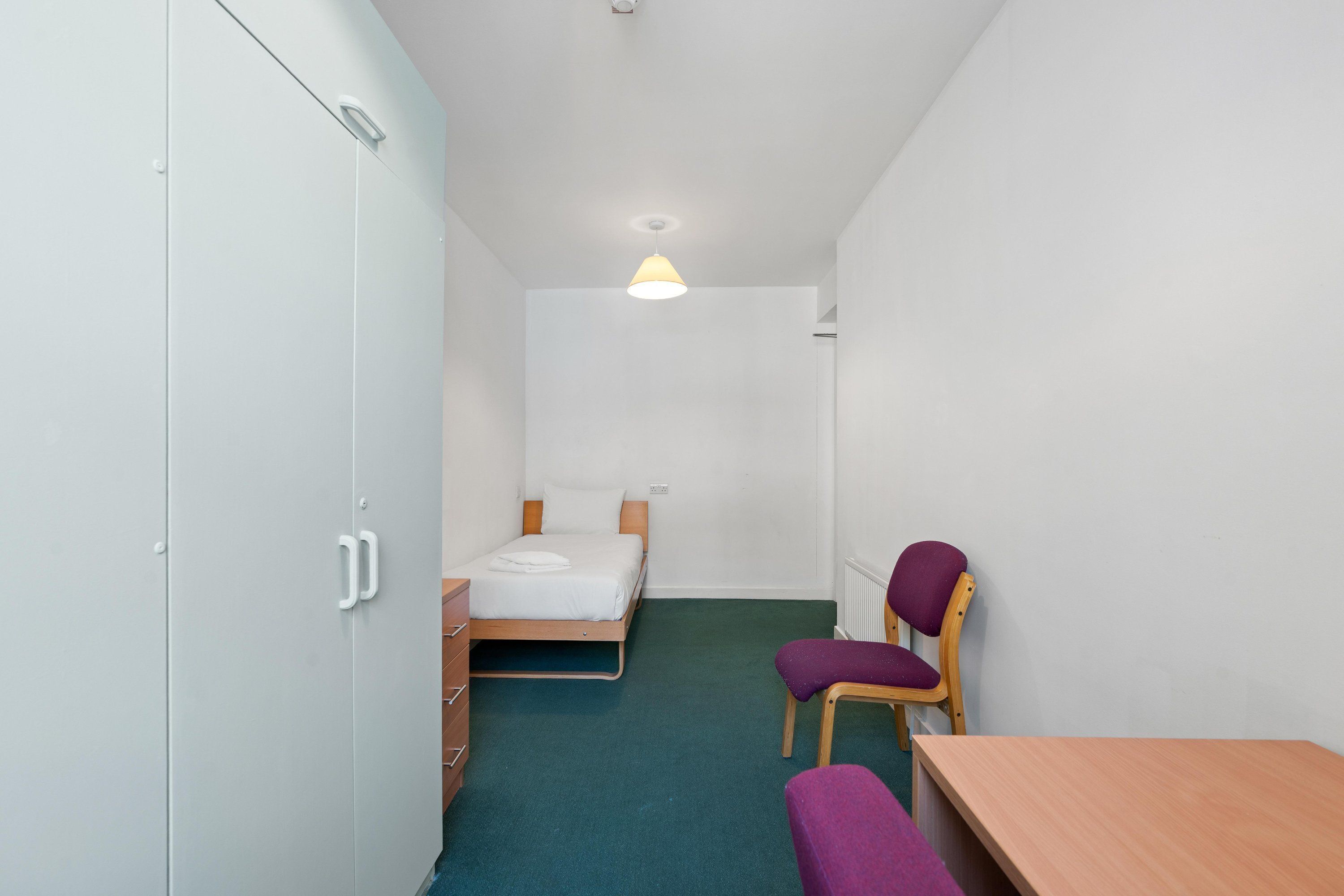 A small, narrow room with a bed, a chair, a table, and a wardrobe.