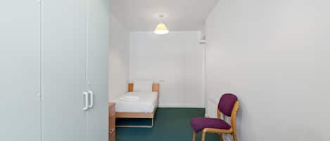 A small, narrow room with a bed, a chair, a table, and a wardrobe.
