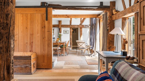 Un interior rústico con vigas de madera a la vista, una zona de comedor con mesa y sillas, y un rincón acogedor con sofá y lámpara.