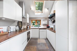 A compact kitchen with white cabinetry, a wooden countertop, a built-in dishwasher, and a window providing natural light. - Medleys Barn By My Getaways (Uckfield)