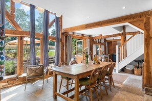 A spacious dining area with a wooden table and chairs, large windows, and a staircase leading to an upper level. - Medleys Barn By My Getaways (Uckfield)