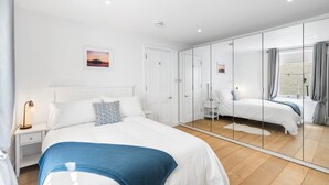 A modern bedroom with a large mirror, a bed with white bedding, a bedside table with a lamp, and a small bedside table with a decorative item. - Bloomsbury Snug By My Getaways (Brighton)